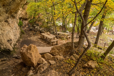 Stone tables and benches are located near a cliff with rock overhangs and alcoves.