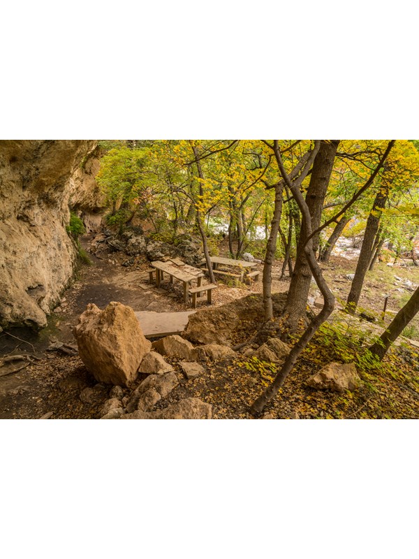 Stone tables and benches are located near a cliff with rock overhangs and alcoves.