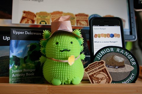 A toy axolotl with a paper ranger hat/badge next to Upper Delaware Junior ranger badge, patch, book