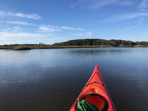Front of kayak surrounded by water, overlooking salt marsh and vegetated island with clear sky above