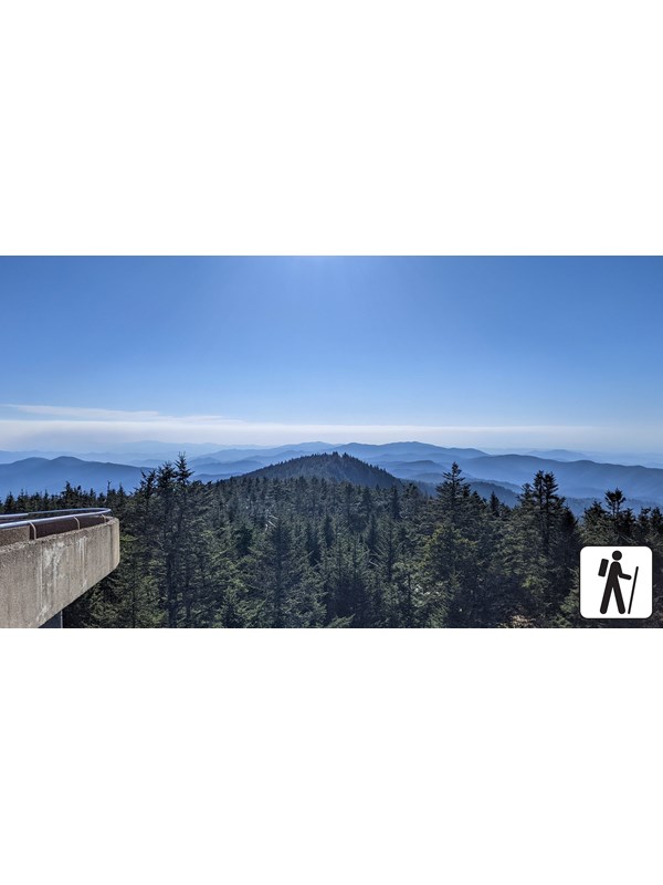An elevated, paved walkway to a round observation tower overlooking mountains. Hiker icon in corner.