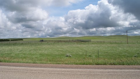 View from a highway into the prairie; in the foreground a rectangular fence breaks up a field.