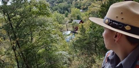 a female ranger in flat hat and uniform on overlook with red mill and blue spring below