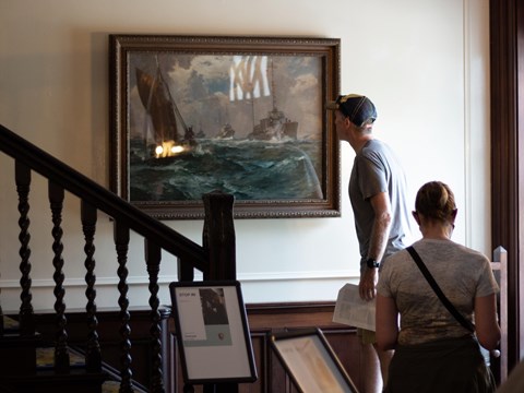 A man standing on a stair landing is looking closely at a naval painting.