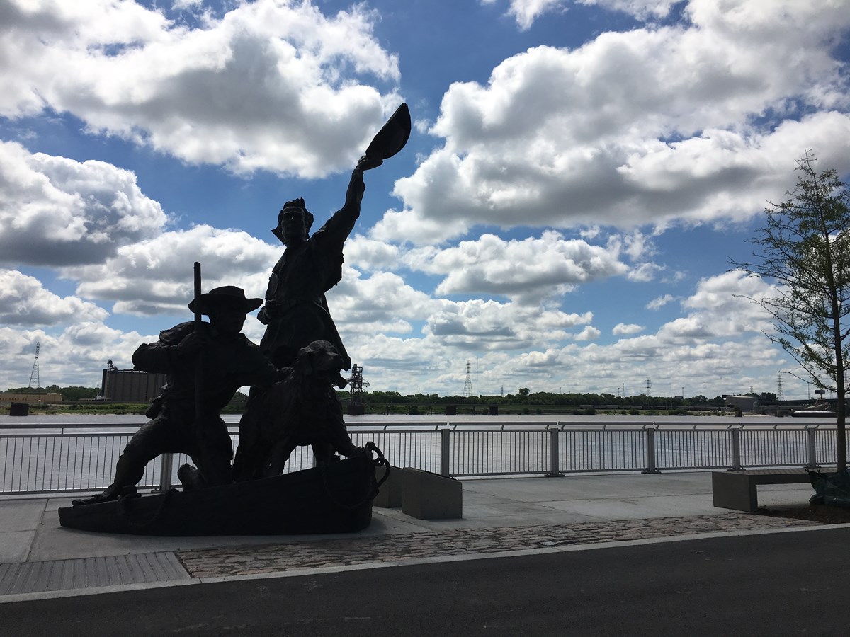 Image of a large statue of two men and a dog silhouetted against a blue sky with many clouds