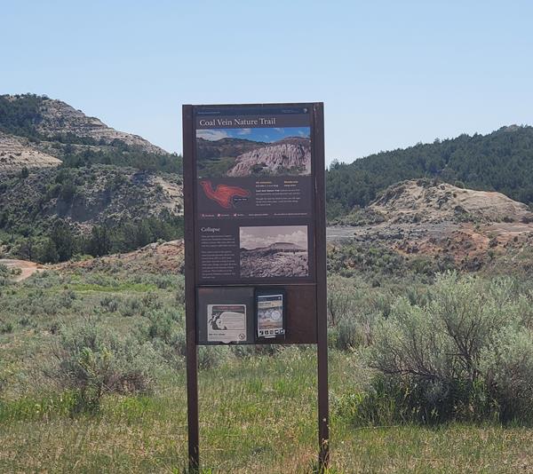 A trailhead sign. Behind it, buttes rise against a blue sky, and grass and sage grow on the ground.