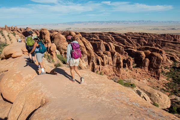 Three hikers walk on a narrow fin of sandstone, with more fins behind them