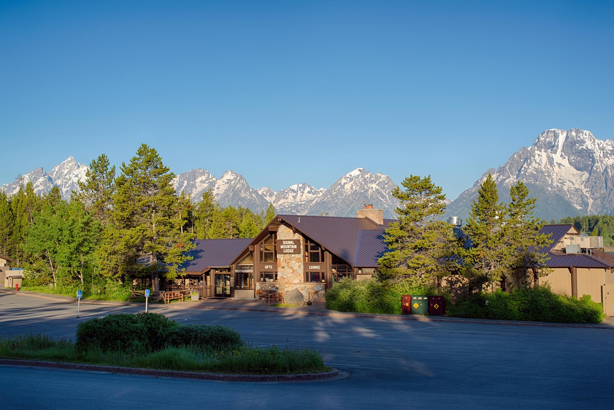 A wide angle image of a wooden lodge with trees and mountains with a blue sky.