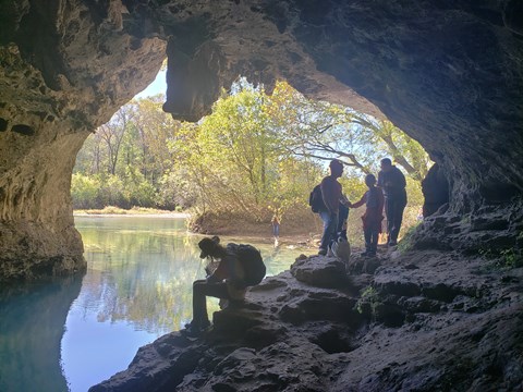looking out of a gray rock cave, blue water in the cave and in the distance, hikers on rocks of cave