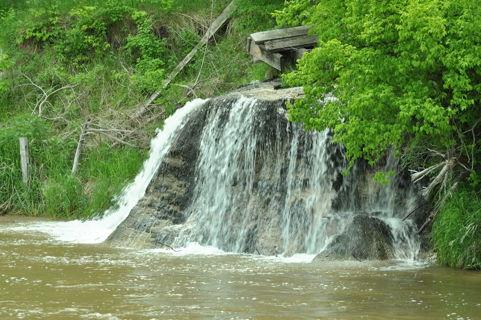 Waterfalls flow into a river with green trees an banks on their perimeter.