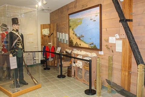 A museum display with manikins in uniforms, a historic painting and a large anchor on the wall.