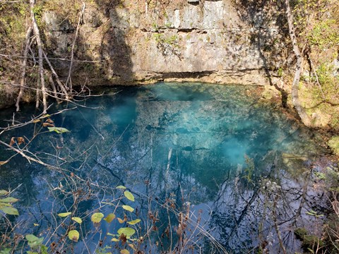 a deep blue spring is at the bottom of a cliff face of grey rock