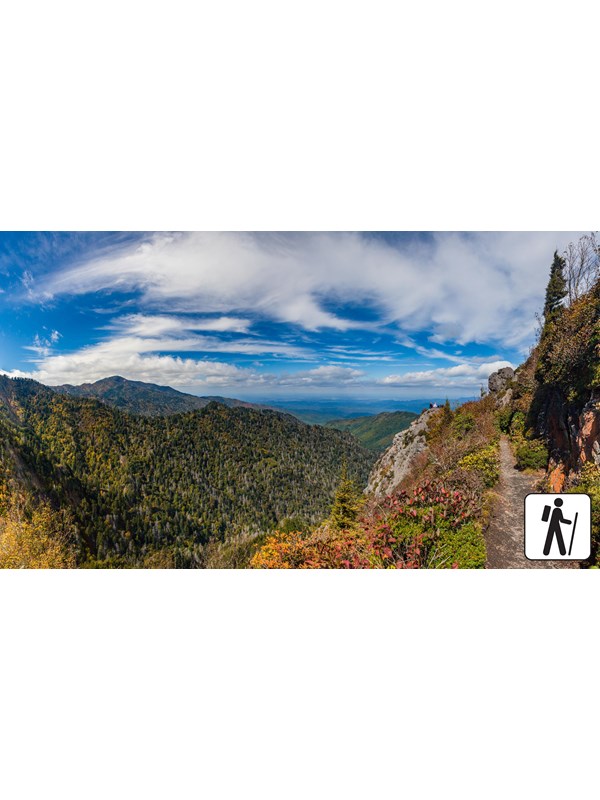 A narrow trail with sweeping views to the left on a partly cloudy day. A hiker icon in corner.