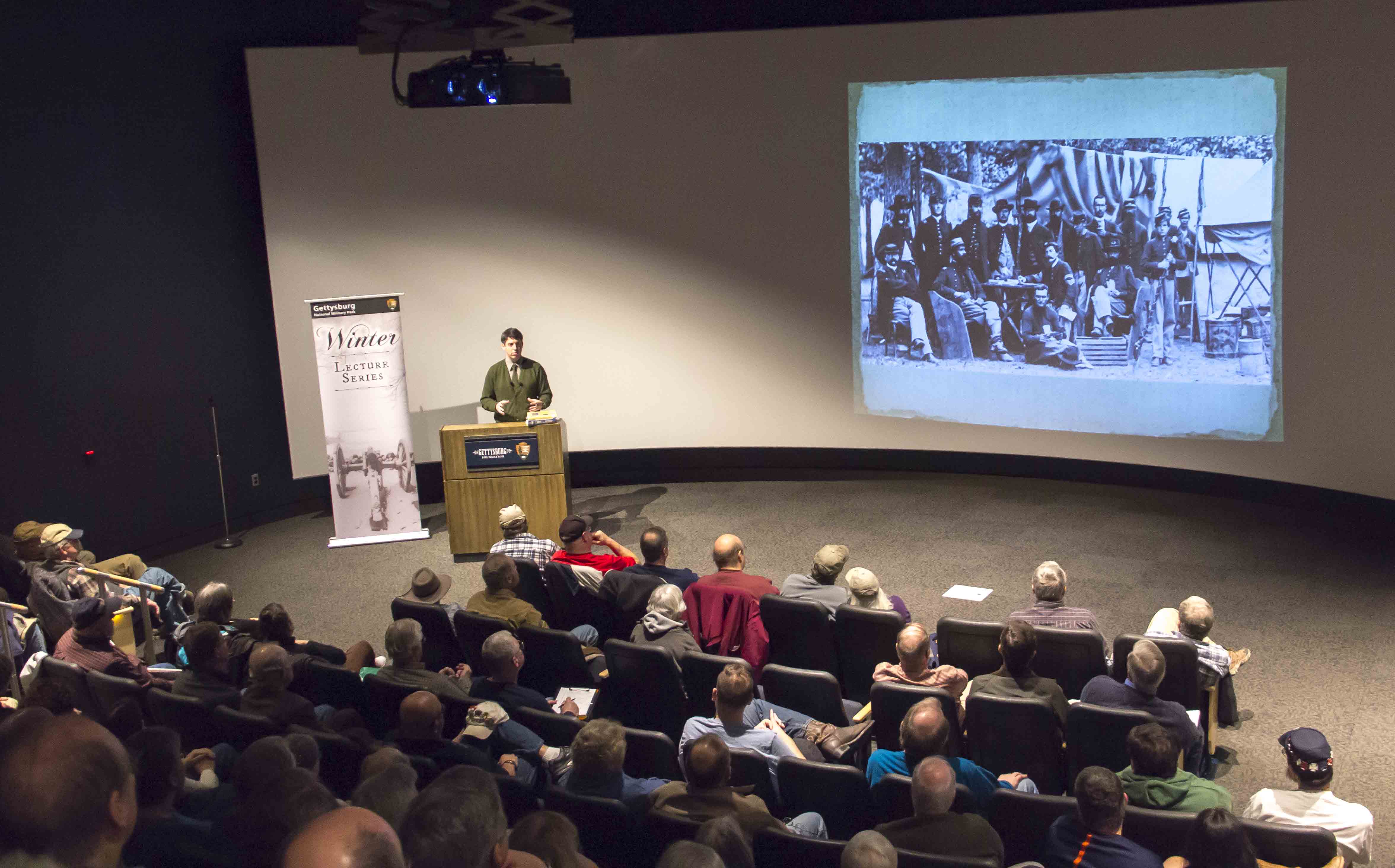 A full theater as seen from the back of the audience. A large white screen, a speaker and podium.