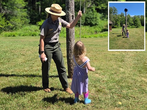 A park ranger stands beside a wooden pole and demonstrates to a small child how to ring the bell.