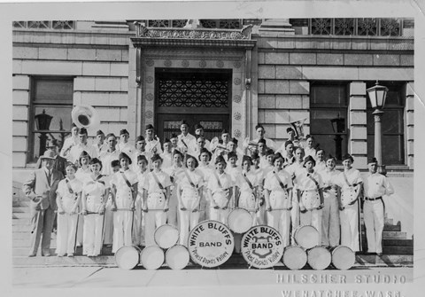 Historic photo of a school band in uniforms standing in front of a brick building