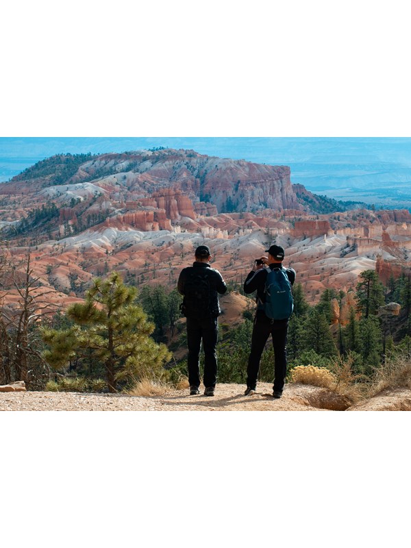 Two hikers pause along a path to view a red rock landscape of badlands and spires at a cliff's edge