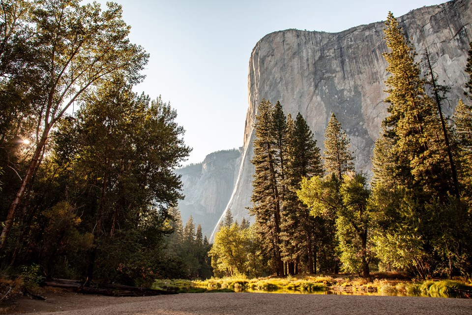 A giant granite monolith looms over trees, a river, and a sandy beach