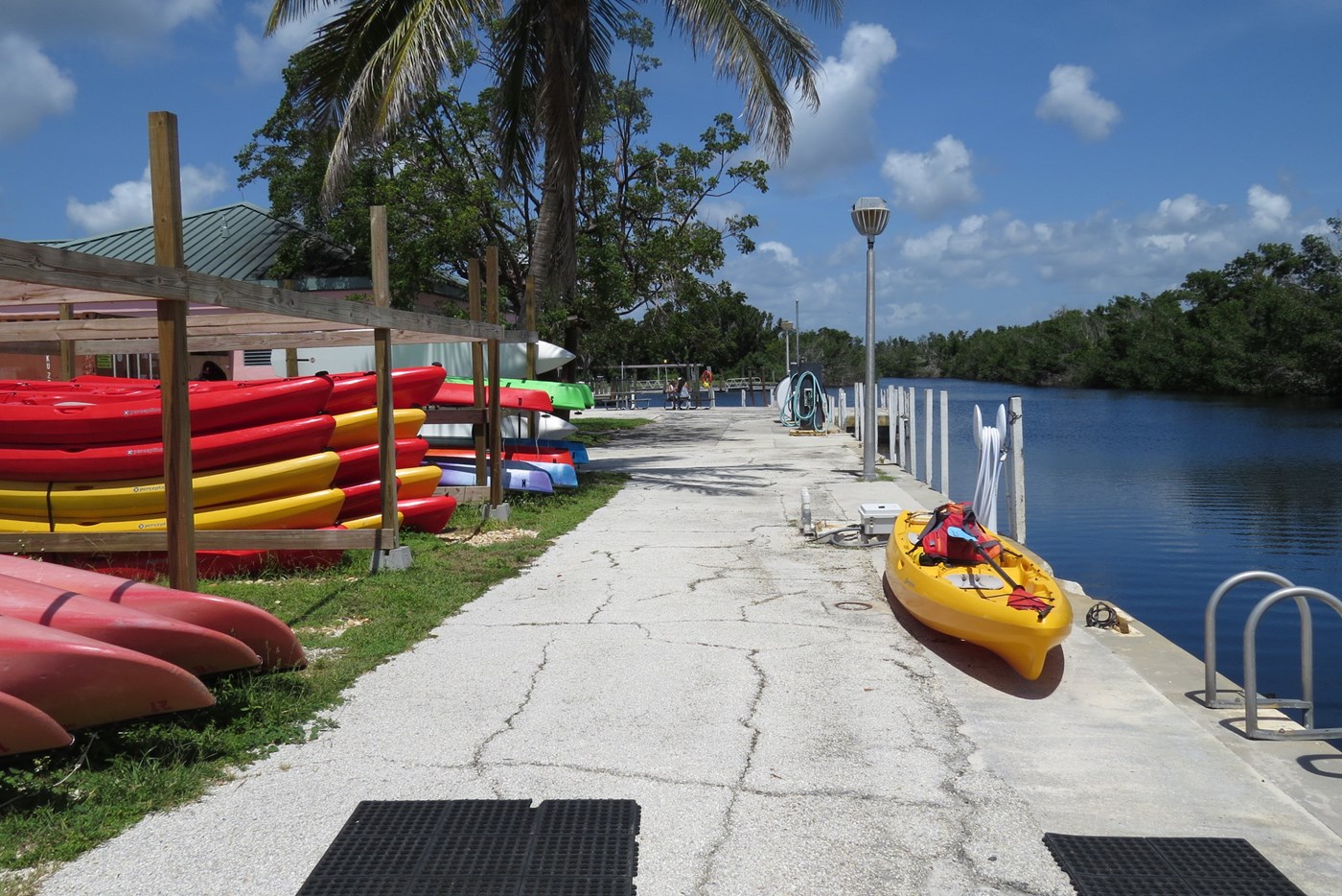 Red and yellow kayaks stacked on the grass beside a cement wall and blue water