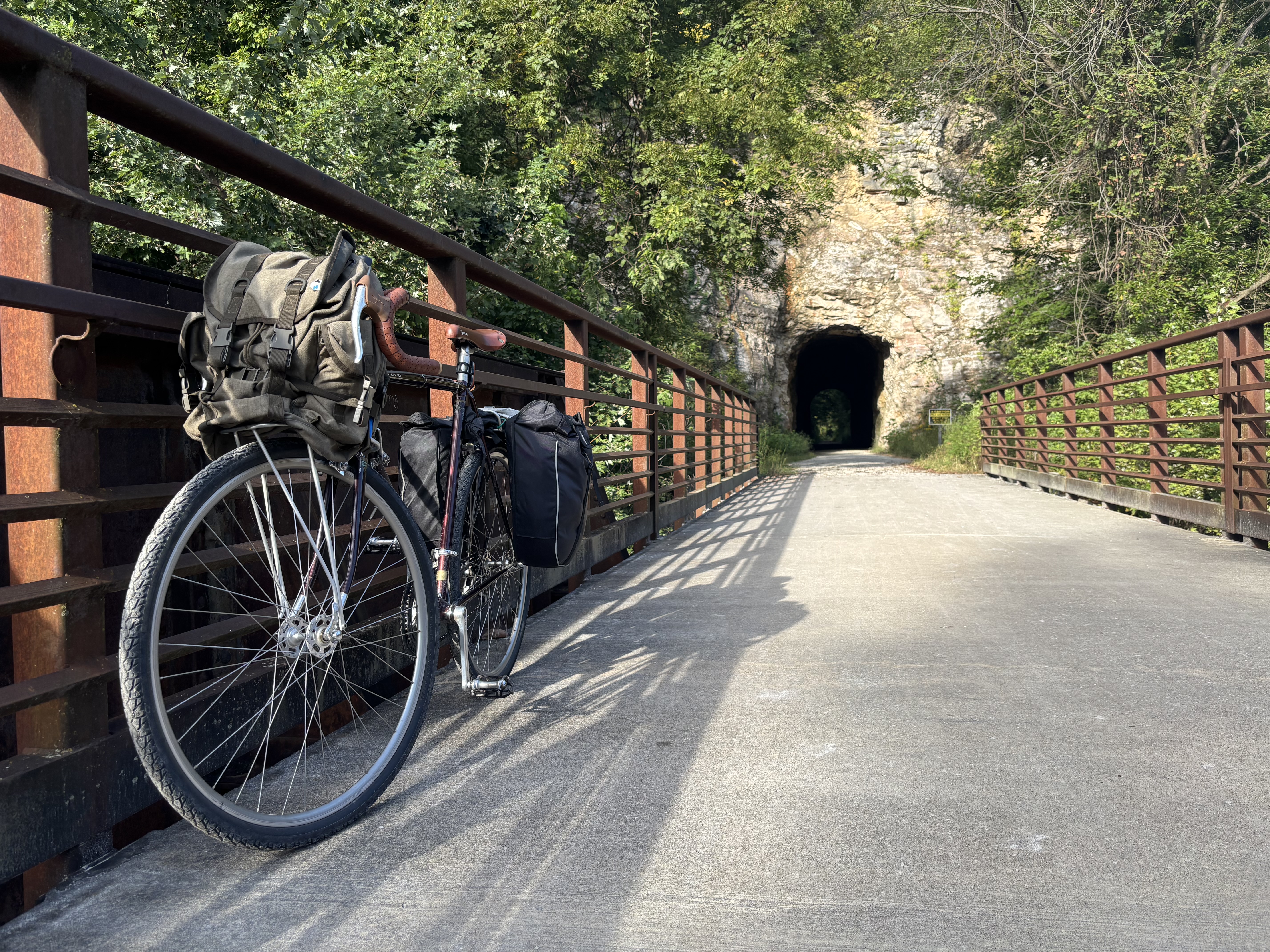 A bike strapped with bags leans on bridge railings with a tunnel through a cliff in background