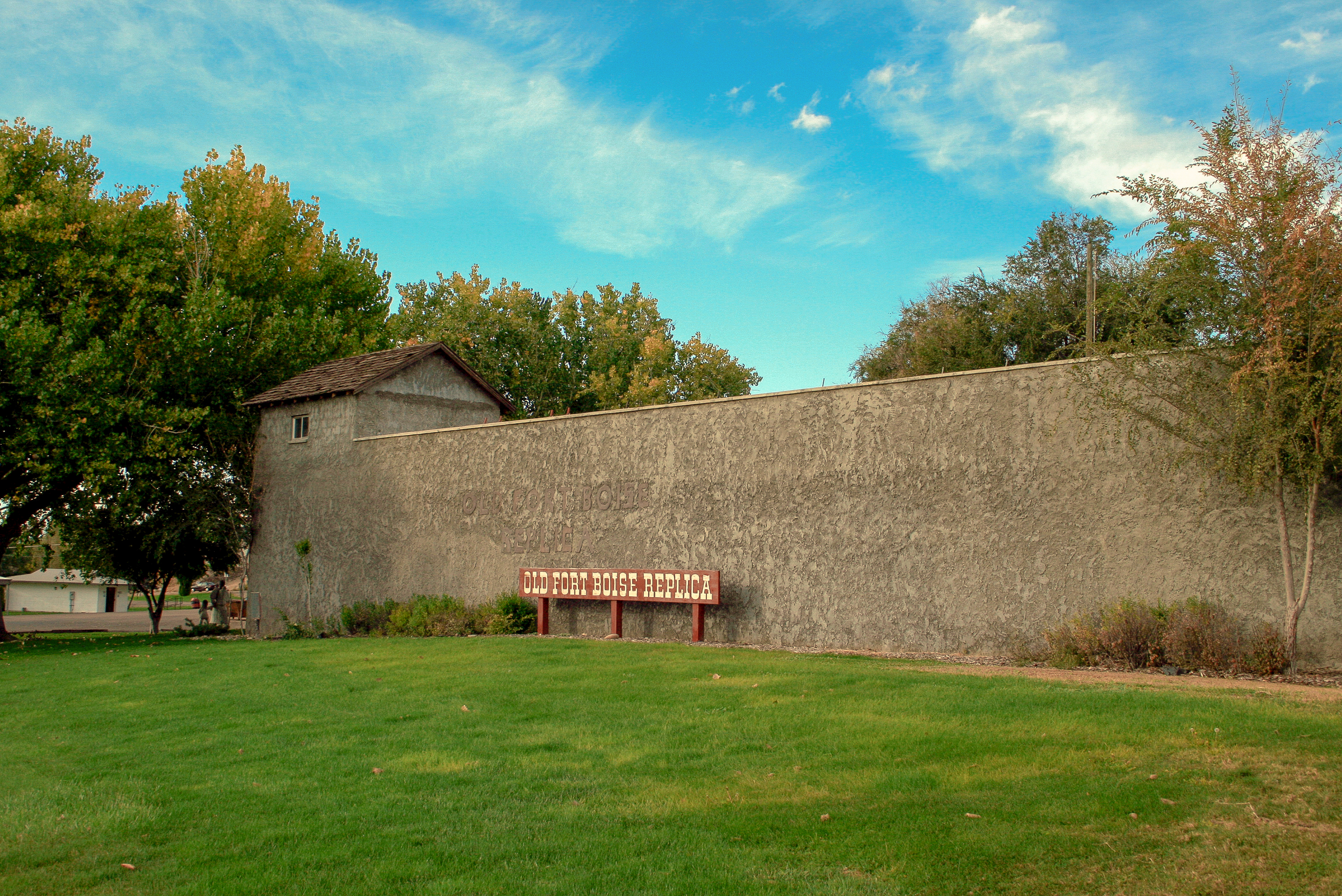 A grey fort wall with a tall corner blockhouse surrounded by green trees and grass under a blue sky.