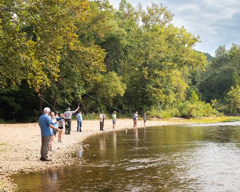 A group of visitors practice casting fly rods along the river's edge.