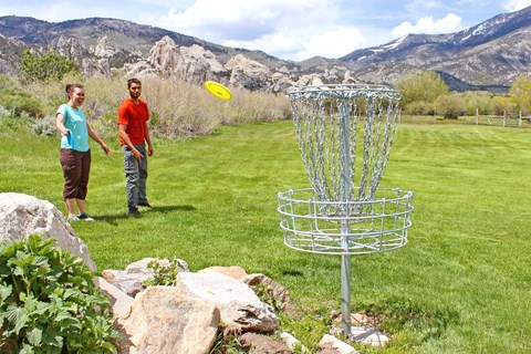 Two people watch a yellow disc flying toward the metal disc golf basket, castle rocks in the back.