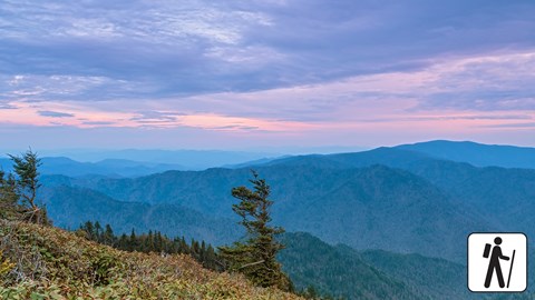 A sweeping mountain vista below a pink and light purple sky. Hiker icon in corner.