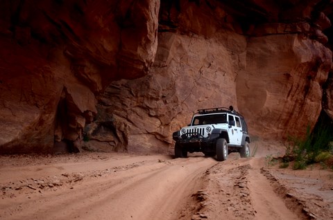 A white jeep traverses a sandy road at the bottom of a red rock canyon.