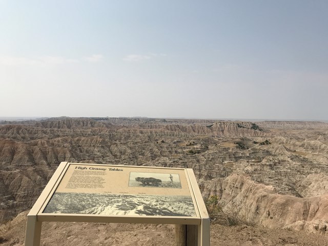 A wayside exhibit sign in the foreground with vast badlands wilderness beyond under blue sky