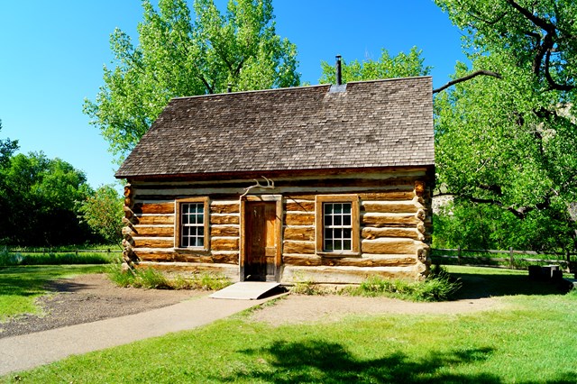 A log cabin in a grassy area with trees and a fence behind it and a sidewalk in front