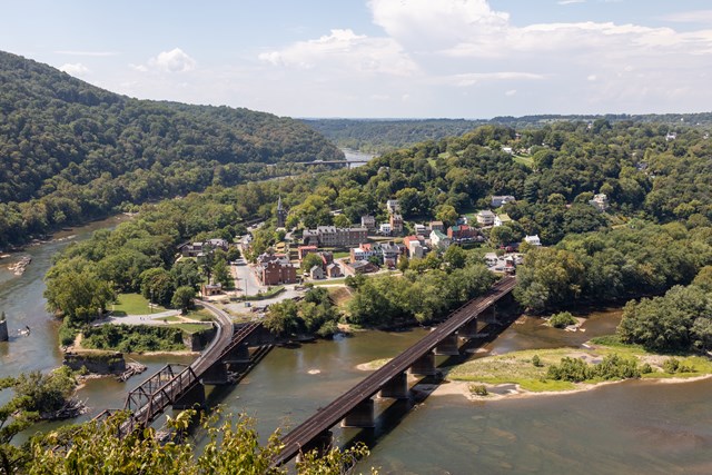 Elevated view over green hills to the town of Harpers Ferry, the rivers below it and two railroad bridges crossing the water