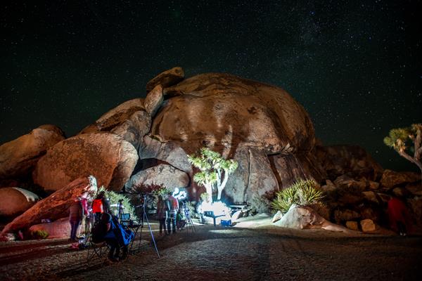 People photographing the night sky over a large rock formation and Joshua trees.