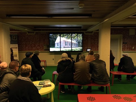 People sitting on benches watching a movie on 3-paneled television screen which shows a wooden house