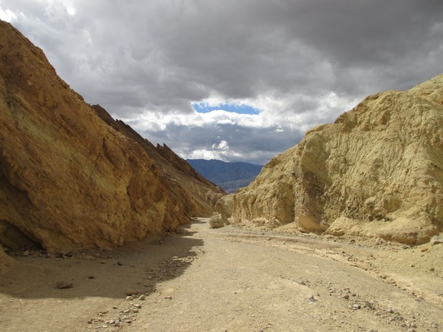 A gravel wash winds between bare yellow hills, with dark clouds overhead.