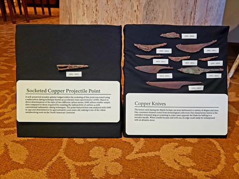 Weathered copper projectile point and knives in a display case.