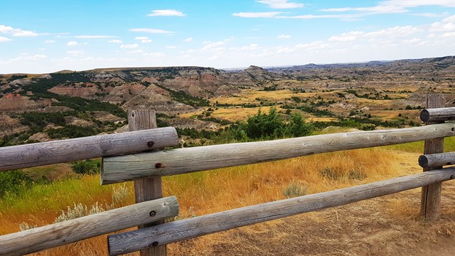 A split-rail log fence in front of a wide view of eroded, layered buttes and golden prairie under a blue sky