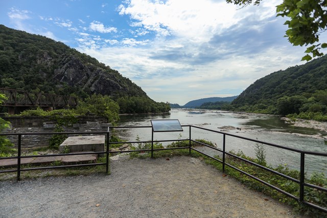 Gravel overlook with a black metal railing and an information panel above a wide river lined with wooded mountains and a truss railroad bridge