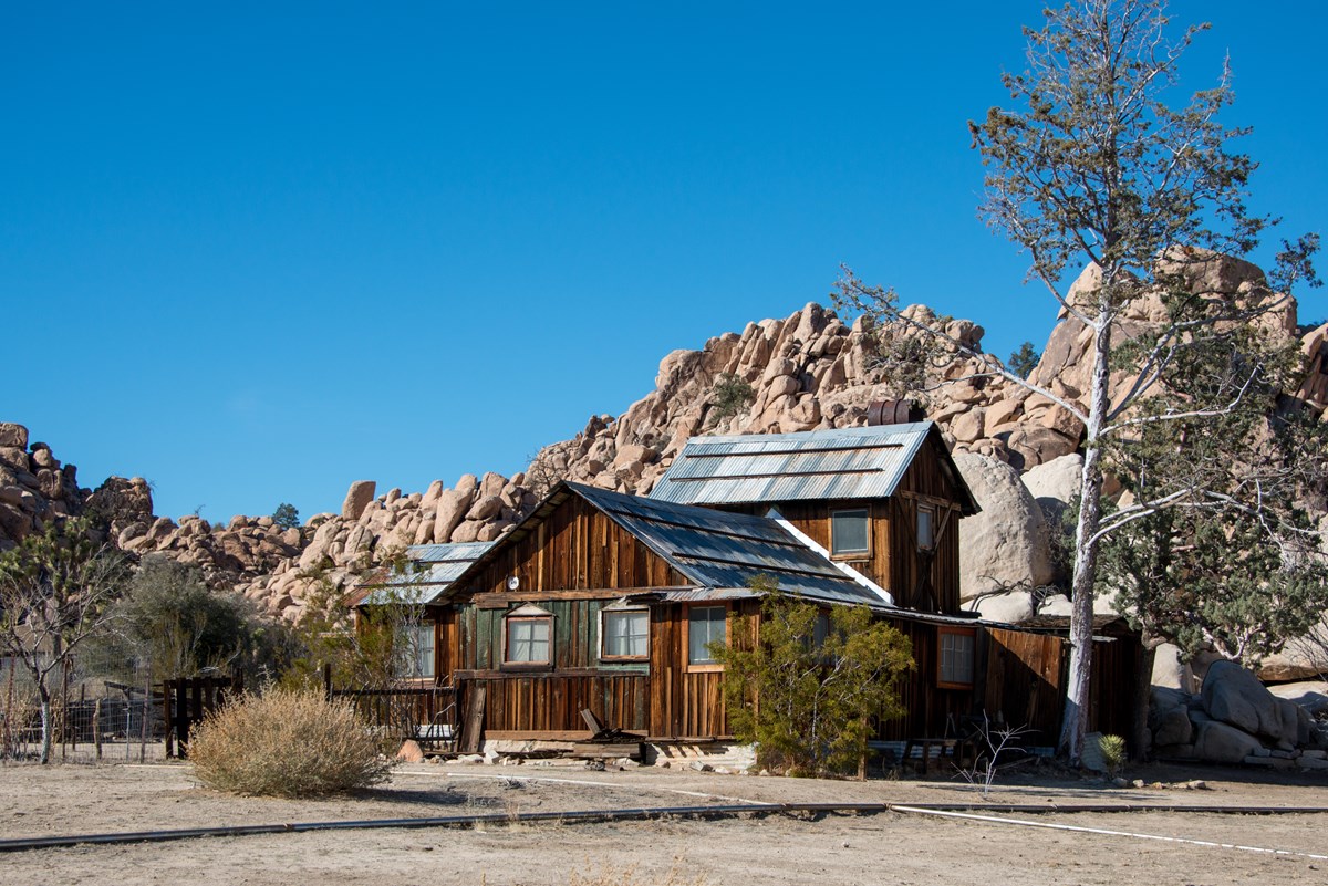 An old wooden two story ranch house with a metal roof in front of a large rock formation.