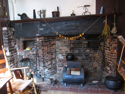 A wooden chair next to a brick fireplace with a wood mantle that has cups on top