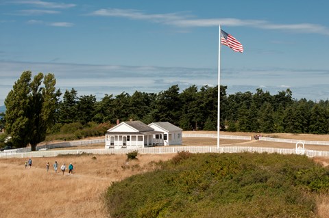 Historic white buildings in a grassy field. Hikers are going along various trails.