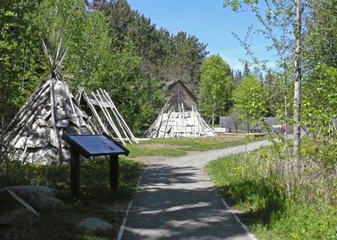 Bark dwellings along a gravel path in front of a reconstructed historic wooden building.