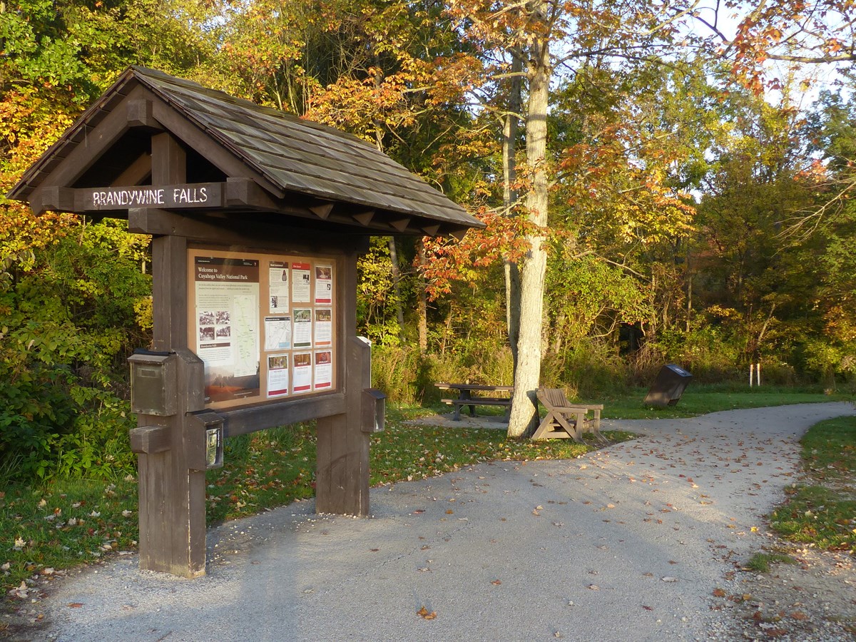 A roofed Brandywine Falls information kiosk stands along a paved path past picnic tables.