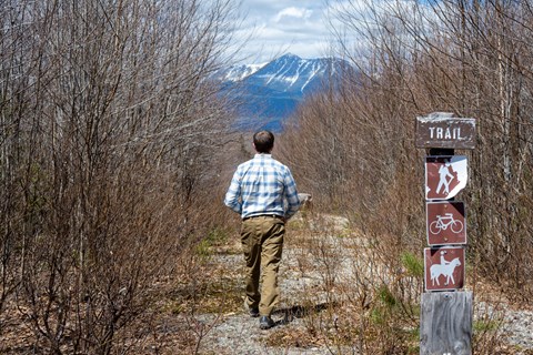A hiker at the gravel trailhead area with a view of Katahdin, a tall mountain in the distance.