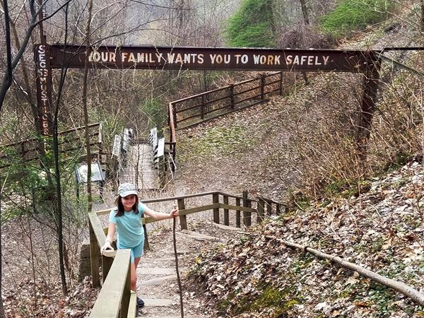 A young hiker on wooden stairs. A sign above her reads, "Your Family wants you to work safely"