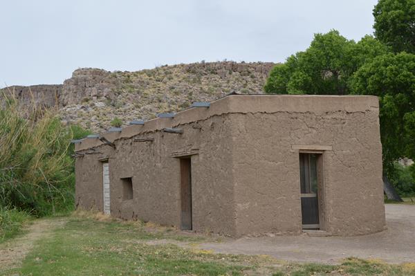 An small adobe building sits in a flat near cottonwood trees and limestone cliffs.