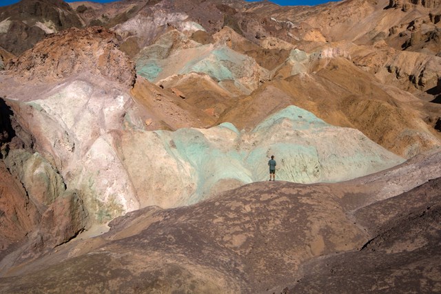 A person stands in front of hills colored brown, cream, pinkish and greenish