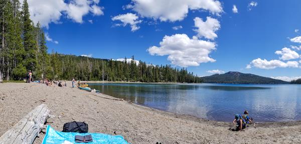People on a gravel beach on the shore of a mountain lake lined by conifers and a large volcanic peak