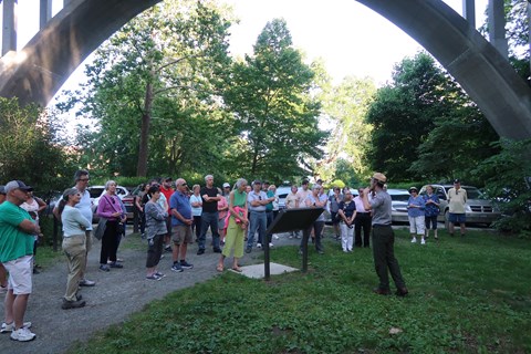 Group of visitors listen to Ranger talking with large cement bridge in background