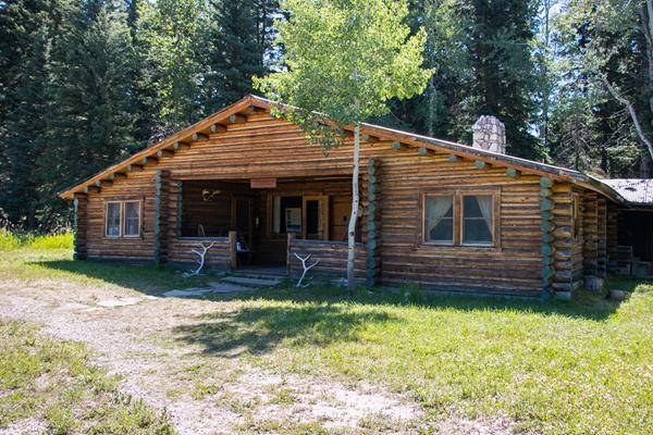 A brown, log cabin building with a river stone chimney and elk antlers resting on the front porch.
